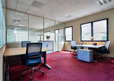 Bureau spacieux avec postes partagés et espace privatif, moquette rouge, mobilier ergonomique et cloison vitrée laissant entrer la lumière naturelle.