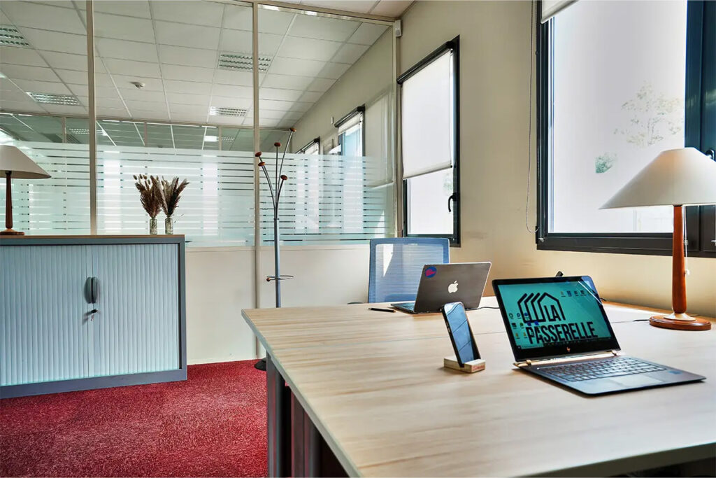 Bureau lumineux avec grands postes de travail, ordinateurs portables, fauteuils ergonomiques, logo La Passerelle affiché sur écran.