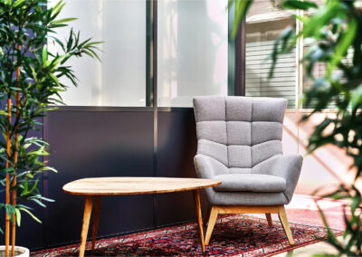 Coin attente avec fauteuil design gris capitonné, table basse en bois et tapis oriental, entouré de plantes vertes.