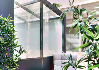 Coin détente avec fauteuil gris capitonné et table basse en bois, baigné de lumière naturelle grâce à une grande verrière et décoré de plantes.