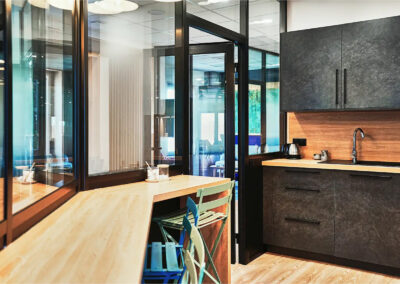 Cuisine équipée avec mobilier moderne noir et bois, espace bar avec chaises hautes colorées et grande verrière donnant sur l’espace commun.