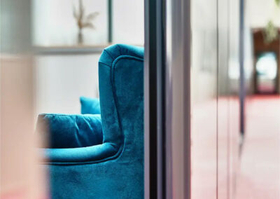 Gros plan sur un fauteuil bleu en velours placé dans le couloir vitré avec moquette rouge, ambiance élégante et moderne.