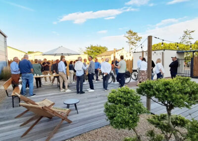 Événement extérieur rassemblant des participants sur la terrasse en bois de La Passerelle, avec tables, bancs et parasols sous un ciel ensoleillé.