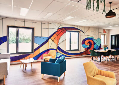 Salon commun avec fresque murale colorée, fauteuils jaunes et bleus, grandes fenêtres et table de réunion en bois.