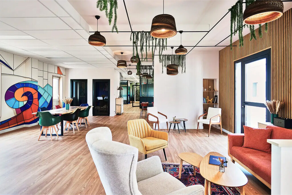 Grand espace de vie avec table en bois massif, chaises colorées, suspensions lumineuses design et cuisine ouverte, ambiance chaleureuse et végétalisée.