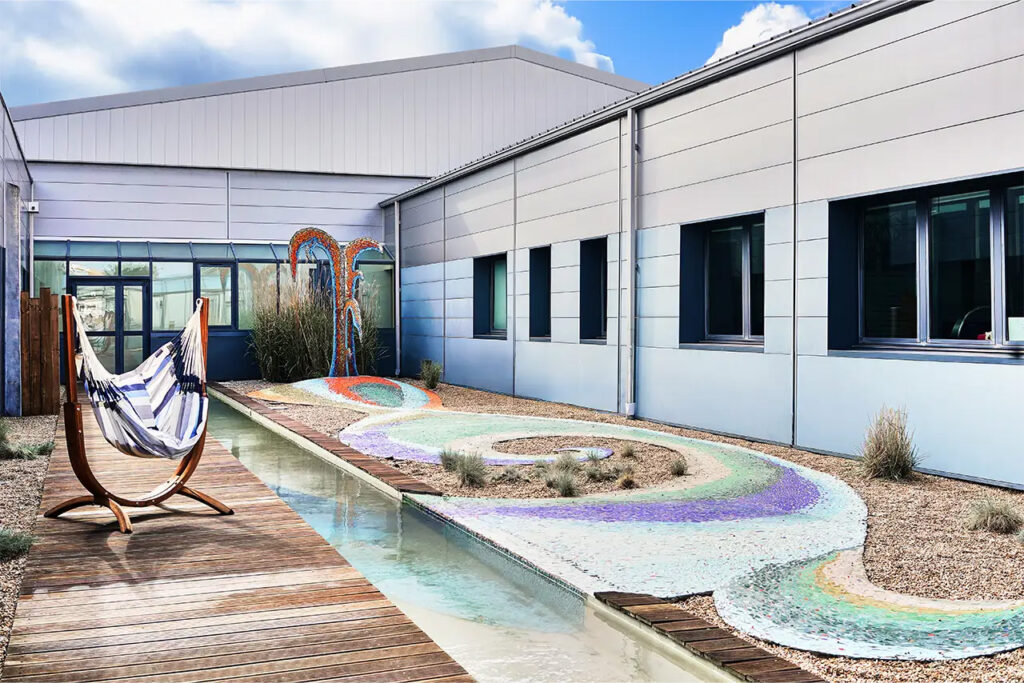 Patio aménagé avec hamac suspendu, allée en bois, bassin d’eau et mosaïque colorée au sol.