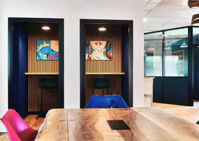 Phone box insonorisée avec assise haute, parois vitrées et décoration murale pop art colorée représentant une femme mettant son doigt devant sa bouche, idéale pour appels confidentiels.