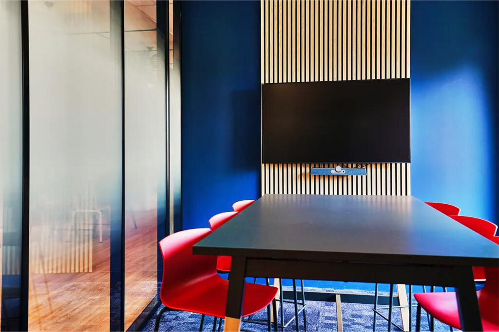 Salle de réunion Adrien avec table haute noire rectangulaire, chaises rouges design et écran mural fixé sur un décor bois et bleu.