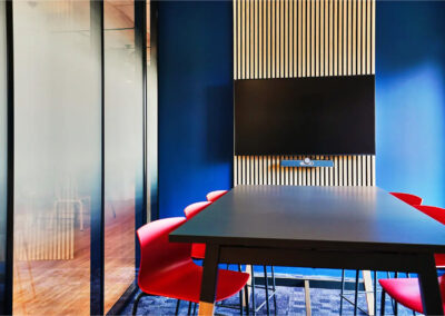 Salle de réunion Adrien avec table haute noire rectangulaire, chaises rouges design et écran mural fixé sur un décor bois et bleu.
