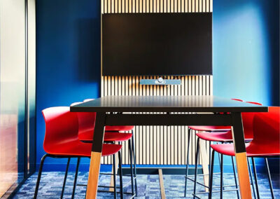 Salle Adrien équipée d’une grande table noire, chaises rouges contemporaines et écran mural, entourée de parois vitrées et d’une ambiance professionnelle.