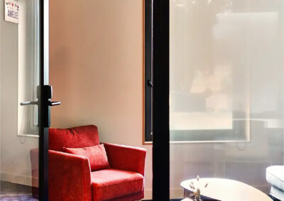 Salle de réunion Amélie aménagée avec fauteuil rouge et canapé gris, tables basses en bois et lumière naturelle donnant sur l’extérieur.