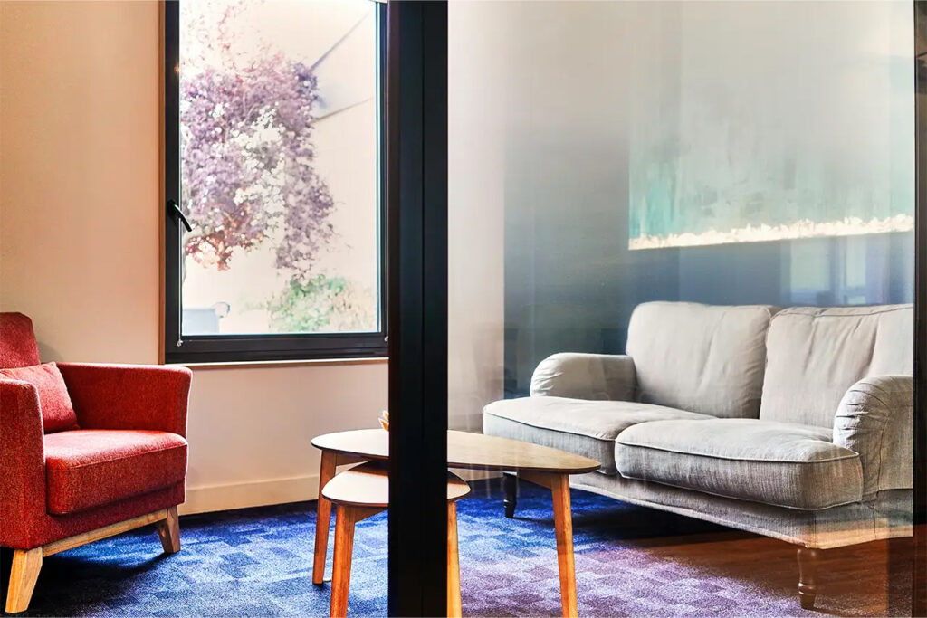 Espace de réunion Amélie avec fauteuil rouge, canapé gris et tables basses en bois, décor chaleureux et ambiance intimiste.