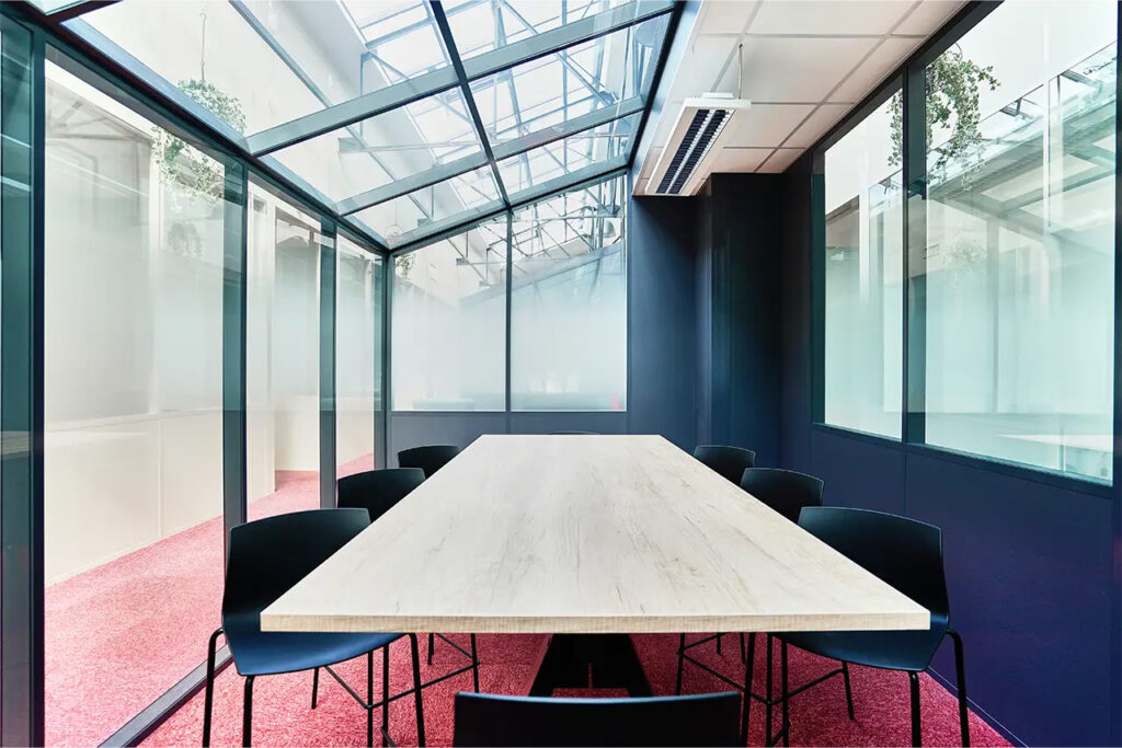Salle de réunion Benoît avec grande table en bois clair, huit chaises noires et parois vitrées sous verrière, idéale pour réunions d’équipe et ateliers collaboratifs.