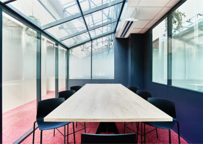 Salle de réunion Benoît avec grande table en bois clair, huit chaises noires et parois vitrées sous verrière, idéale pour réunions d’équipe et ateliers collaboratifs.