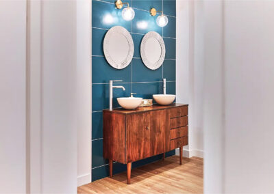 Salle de bain élégante avec meuble en bois rétro, deux vasques modernes, miroirs ronds décoratifs et carrelage mural bleu.