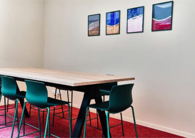 Salle Patricia avec grande table en bois, chaises vertes design et mur décoré de tableaux colorés, parfaite pour les échanges d’équipe.