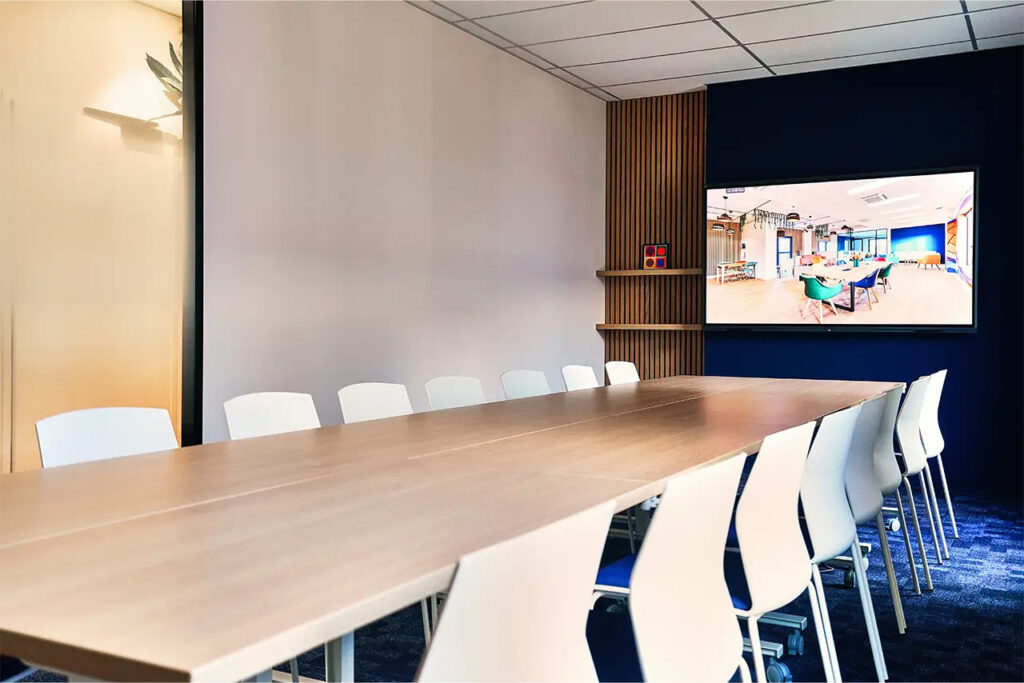 Grande salle de réunion Pio pour 16 personnes avec table en bois clair, chaises blanches et écran connecté fixé sur un mur bleu.