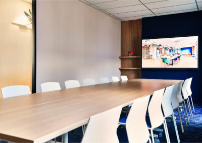 Grande salle de réunion Pio pour 16 personnes avec table en bois clair, chaises blanches et écran connecté fixé sur un mur bleu.