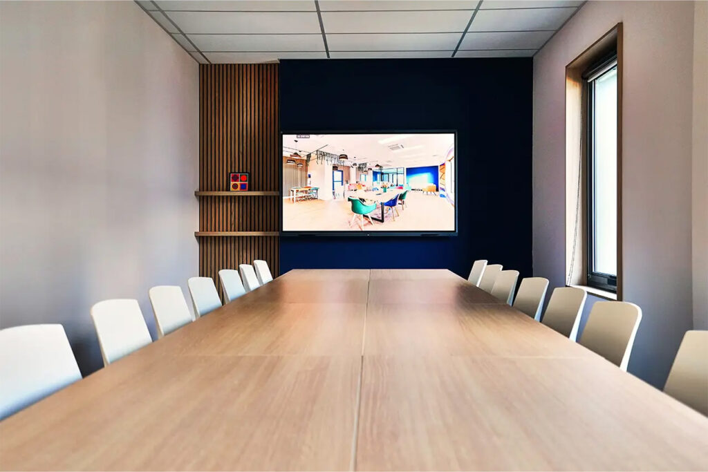 Salle Pio avec grande table de réunion, chaises blanches et écran mural connecté, décorée de lames bois et d’un mur bleu.