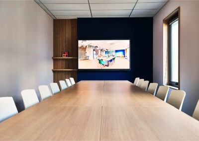 Salle Pio avec grande table de réunion, chaises blanches et écran mural connecté, décorée de lames bois et d’un mur bleu.
