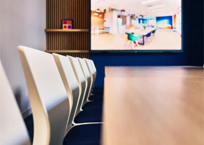 Grande salle de réunion Pio avec table en bois clair, chaises blanches et écran connecté fixé sur un mur bleu.