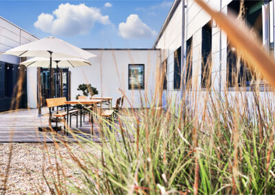Terrasse aménagée avec tables en bois, bancs, parasols blancs et sol en bois, parfaite pour déjeuner ou travailler en extérieur.