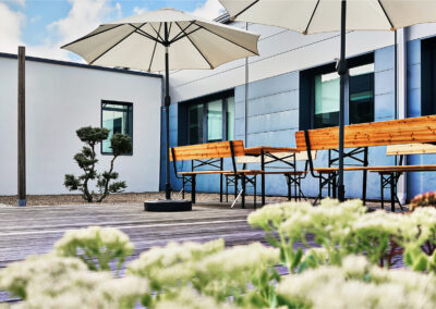 Terrasse en bois équipée de bancs, tables et parasols, entourée de végétation et baignée de lumière naturelle.