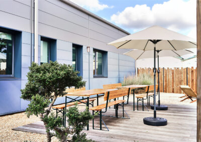 Terrasse moderne avec bancs en bois, grands parasols blancs et plantations décoratives autour de l’espace extérieur.