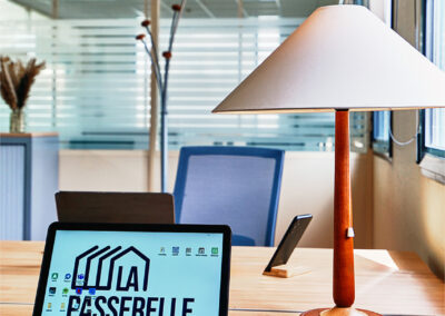 Bureau en bois clair avec ordinateur portable affichant le logo La Passerelle, lampe design et assises ergonomiques.