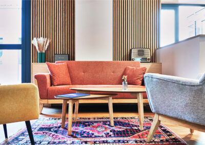 Salon moderne avec canapé rouge, fauteuils gris et jaunes, tables basses en bois et tapis décoratif coloré.